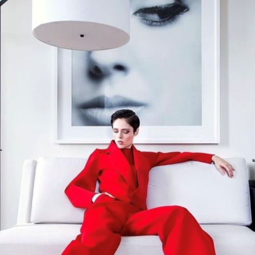 Woman in a red suit seated on a white couch beneath a large black‑and‑white portrait, a bold composition showcasing contrast and style, with contemporary figurative canvas prints available through Wilde Portraits.